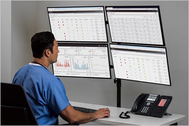 Clinician in scrubs sitting at the Masimo SafetyNet command center with four computer screens.