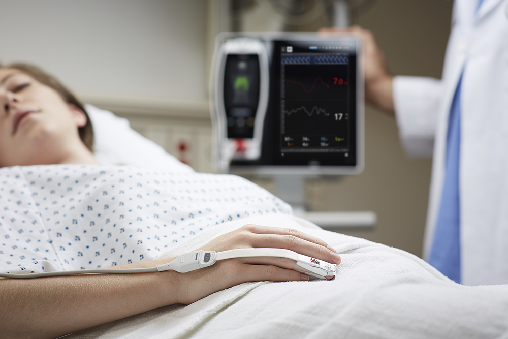 Patient sleeping in hospital bed with Masimo monitor on finger and Masimo Root device in background.